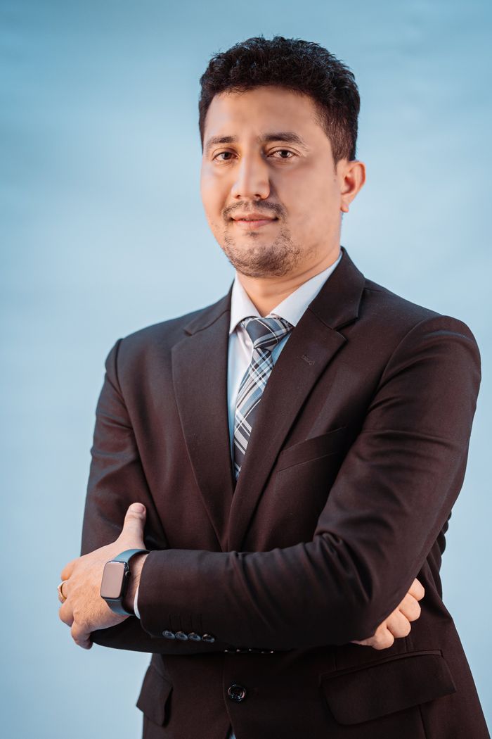 A confident businessman posing with arms crossed wearing a formal suit against a blue background.